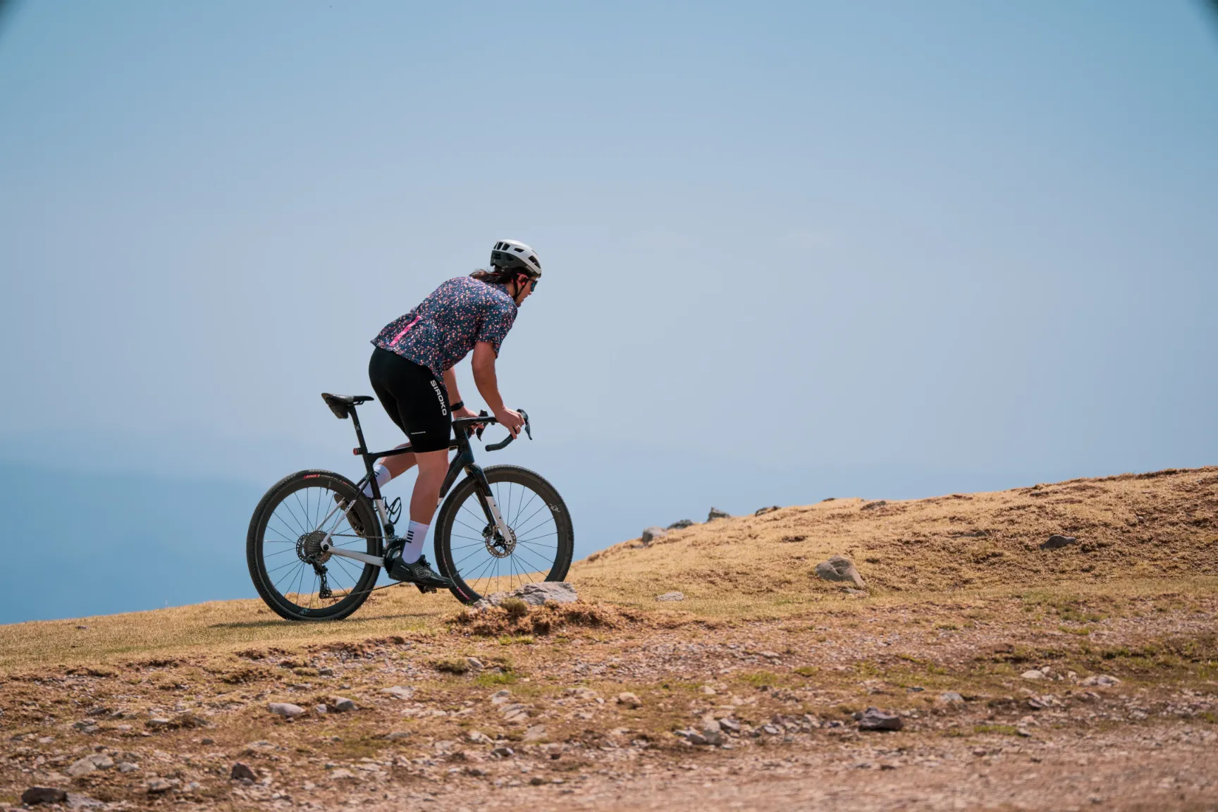 Rider de La Fuga × UNÎCA subiendo una loma de gravel sobre cielo azul, vista trasera